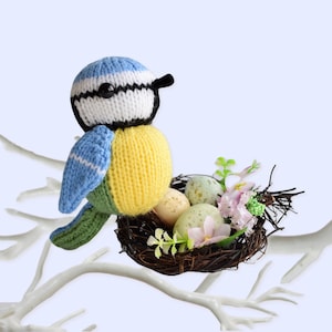 Może przedstawiać: Szydełkowany ptak modraszka z biało-niebieską głową w paski, w pobliżu gniazda. Ptak ma żółte ciało, niebieskie skrzydła i zielony ogon. Gniazdo zawiera nakrapiane jaja, różowe kwiaty i zielone liście.