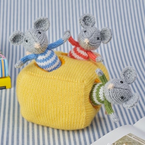 May include: Three small, grey, crocheted mice figures emerging from a large, yellow, crocheted block. One mouse wears a blue and white striped jumper, another a red and white striped jumper, and the third a green and white striped jumper.