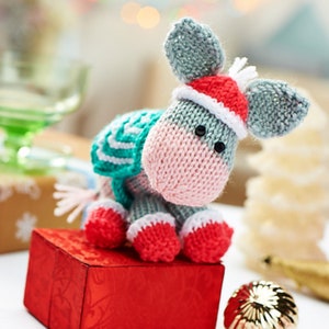 May include: A knitted grey donkey wearing a red and white Santa hat and a teal and white striped scarf. The donkey is sitting on a red gift box.