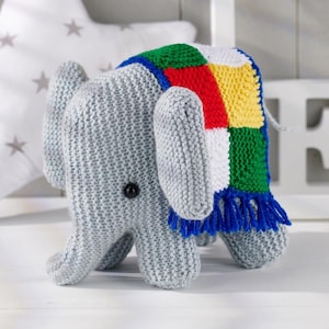 Pode incluir: Um elefante de brinquedo em crochê cinza com uma pequena manta sobre as costas. A manta é composta por uma grade de quadrados verdes, vermelhos, brancos e amarelos, finalizada com uma franja de fio azul na borda inferior. O elefante tem olhos pretos.