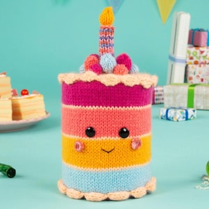 May include: A cylindrical knitted birthday cake toy featuring horizontal stripes of magenta, orange, yellow, and light blue. It has a smiling face with black bead eyes, pink cheeks, and a single knitted candle on top with a yellow flame.