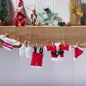 Könnte beinhalten: Eine festliche Weihnachtsdekoration mit Miniatur-Strickkleidung und Ornamenten. Unter einem Regal mit einem Weihnachtsbaum, einer Mäusefigur und einem Geschenk hängt eine Schnur mit winzigen Weihnachtsmann-Anzügen, Mützen und Unterwäsche.