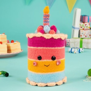 May include: A knitted birthday cake with a smiling face, featuring layers of pink, orange, yellow, and blue. It has a candle on top and is decorated with colorful knitted balls. The cake is approximately 6 inches tall.