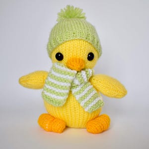 Op de afbeelding: Een geel gehaakt eendje met een lichtgroene gebreide muts met een pompon bovenop. Het eendje draagt een sjaal met horizontale lichtgroene en witte strepen. Het heeft twee zwarte kraaloogjes en een oranje snavel en oranje voetjes.
