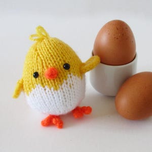 May include: A knitted yellow and white chick toy with black eyes, an orange beak, and orange feet, positioned next to a white ceramic egg cup containing a brown egg and a second brown egg resting on the surface beside the cup.