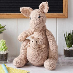 Puede incluir: Un peluche de canguro tejido en color beige claro que incluye un pequeño canguro bebé dentro de su bolsa frontal. El canguro tiene dos ojos oscuros, orejas puntiagudas y una textura de hilo. Es una figura de peluche suave en posición sentada.