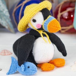 Könnte beinhalten: Ein handgestrickter Papageitaucher-Vogel mit gelbem Hut, blau-orangem Schnabel und orangefarbenen Füßen. Er hält einen blauen Strickfisch. Der Papageitaucher hat eine weiße Brust und schwarze Flügel und Rücken.