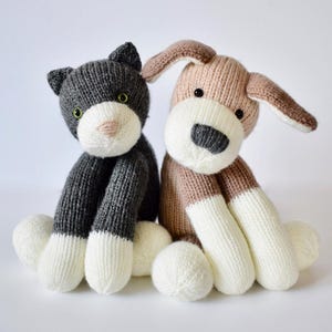 May include: A pair of knitted plush toys consisting of a dark gray cat with a white muzzle and paws, and a light brown dog with a white muzzle, chest, and paws. Both stuffed animals feature black button eyes and stitched nose details.