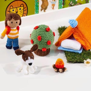 May include: A knitted playset featuring a doll with brown hair, a white and yellow striped shirt, and blue pants, a brown and white dog, a green tree with red spots, an orange tent with a blue pom-pom, and a small campfire on a green patch.