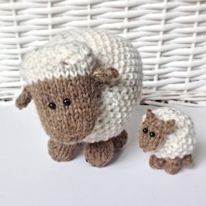 May include: Two hand-knitted sheep figures, one large and one small, featuring cream-coloured bodies and brown faces with small black bead eyes. The sheep have textured, rounded bodies and four small brown legs, crafted with visible yarn stitches.