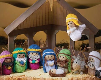 May include: A knitted nativity set featuring a wooden stable with a gold star on top. The set includes figures of people in colorful robes, a small baby in a manger, an angel, a donkey, and a sheep, all arranged on a bed of straw-like material.