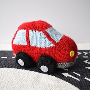以下が含まれることがあります： 鮮やかな赤色で水色の窓が付いた、ニット製のソフトな車のおもちゃ。車両には白いボタンの中心を持つ黒い円形の車輪があり、前面には黄色いボタンのヘッドライトが付いています。底面は灰色のニットの縁取りで仕上げられています。