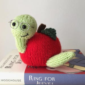 May include: A hand-knitted plush toy shaped like a worm emerging from a bright red apple. The worm is light green with black bead eyes, a stitched smile, and small silver-toned wire glasses. A dark green leaf and brown stem sit atop the red apple.