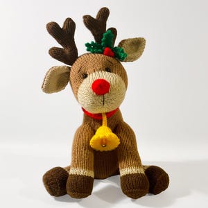 May include: A knitted reindeer plush toy featuring a brown body, a bright red nose, and dark brown antlers. It wears a red collar with a yellow bell attached. A small green holly sprig with a red berry is positioned between the antlers.