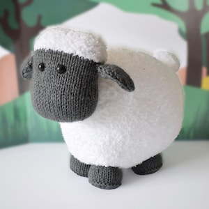 May include: A plush sheep toy featuring a dark gray knitted head and legs with black bead eyes. The body is covered in a white, textured, fuzzy material that mimics wool. The sheep has two dark gray knitted ears extending from the sides of its head.