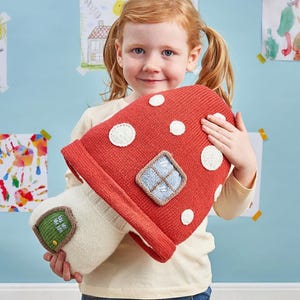 May include: A red mushroom-shaped pillow with white polka dots and a small window design. The pillow has a cream-colored stem with a green door. A child is holding the pillow.