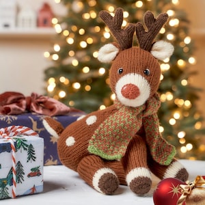 Könnte beinhalten: Ein braunes, gestricktes Rentier mit weißen Flecken, Geweih und roter Nase. Es trägt einen grün-roten Schal. Das Rentier sitzt vor einem Weihnachtsbaum mit Lichtern und verpackten Geschenken.