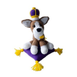 Puede incluir: Un juguete de perro tejido a mano con un cuerpo marrón y blanco, que lleva una corona morada y dorada. El perro está sentado en un cojín morado con borlas amarillas. Tiene una nariz negra y un collar amarillo.
