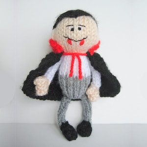 May include: A hand-knitted vampire doll featuring a pale beige face with black hair, a white shirt, grey trousers, and black shoes. The doll wears a black cape with a red collar and a red tie string around the neck, with embroidered facial features.