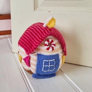 Puede incluir: Un original tope de puerta tejido a mano con forma de casa. El tejado es de color rosa vibrante con rayas rojas, y el cuerpo presenta rayas beige y blancas. Una sección azul representa la puerta, con una ventana blanca. Detalles amarillos y rosas.
