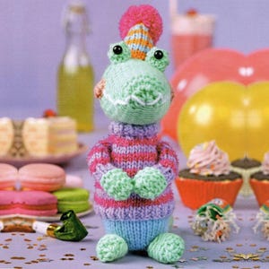 May include: A light green crocheted alligator toy wearing a striped pink and purple sweater and a matching party hat with a pink pom-pom. The toy has black bead eyes and a white stitched mouth, standing upright with small arms and legs.