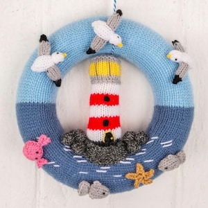 May include: A knitted nautical wreath featuring a lighthouse with red and white stripes, a yellow top, and gray rocks at the base. The wreath is blue with seagulls, a pink seahorse, a starfish, and gray fish.