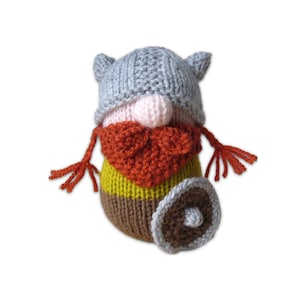 May include: A knitted plush doll shaped like a Viking, featuring a grey helmet, a pale pink nose, a textured orange beard, a yellow and brown body, and a small round brown shield with a grey border held at the side of the body.