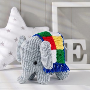 May include: A gray knitted elephant toy with large ears and a colorful patchwork blanket draped over its back. The blanket features squares of red, yellow, green, and white, trimmed with blue fringe. A black button is visible on the elephant's side.