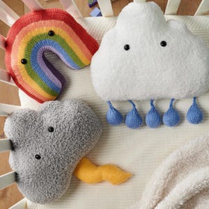 May include: A selection of soft, knitted baby pillows shaped like a rainbow, a cloud with raindrops, and a grey cloud with a yellow accent. The pillows are on a white crib sheet, creating a whimsical nursery theme.