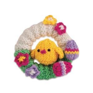 May include: A circular knitted wreath featuring a bright yellow chick in the centre. The wreath is decorated with small knitted flowers in pink, purple, and yellow, along with two patterned Easter eggs in shades of purple, pink, and yellow.