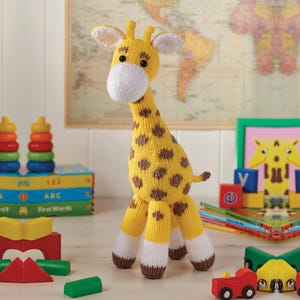Könnte beinhalten: Eine handgestrickte Giraffe in Gelb und Braun mit weißen Akzenten. Die Giraffe hat braune Flecken, eine weiße Schnauze und kleine Ohren. Bunte Holzbausteine und Bücher umgeben das Spielzeug.