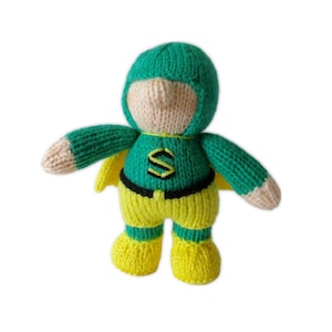 May include: A knitted superhero doll with a green hood, cape, and sleeves, yellow trousers and boots, and a beige face and hands. The doll has a black belt and a yellow and black "S" emblem on the chest.