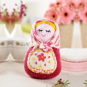 Puede incluir: Muñeca Matrioska tejida a mano en tonos rosa, blanco y amarillo. La muñeca presenta una cara sonriente, un pañuelo en la cabeza y un delantal con un diseño floral. Un encantador ejemplo de artesanía tradicional.
