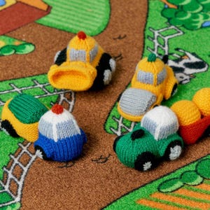 May include: Five knitted toy vehicles in different colors and styles, including a yellow bulldozer, a green car, a blue car, a yellow truck, and a red truck. The toys are on a green and brown patterned surface.
