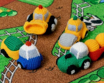 May include: A collection of colorful, knitted toy cars in yellow, green, blue, and red. The cars have black wheels and gray or white accents. Some have small red or green pom-poms on top. The toys are arranged on a brown and green play mat.