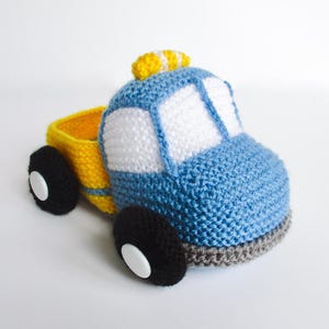 May include: A crocheted toy truck featuring a blue cab with white windows, a yellow truck bed, and four black wheels with white circular buttons at the center. The vehicle has a small yellow textured accent on the roof and a grey base trim.