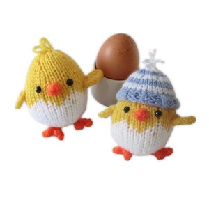 May include: Hand-knitted yellow and white chick egg cosies with orange beaks and feet. One chick wears a blue and white striped hat. A brown egg sits in a white egg cup. These charming cosies add a touch of whimsy to any breakfast table.