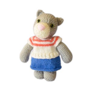 Puede incluir: Muñeca de gato tejida a mano, con pelaje gris, ojos verdes y nariz rosa. El gato lleva una camisa a rayas blancas y coral y una falda azul. La muñeca tiene orejas pequeñas.