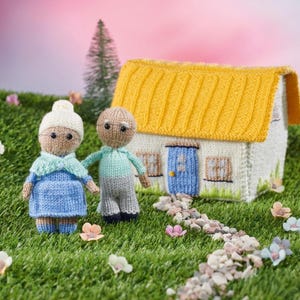 Op de afbeelding: Een gebreide scène met een miniatuur ouder echtpaar en een huis met een geel dak. Het paar draagt blauwe en grijze outfits en het huis heeft een blauwe deur. De scène speelt zich af op een groene, grasachtige ondergrond.