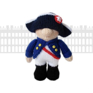 Może przedstawiać: Dzianinowa lalka przedstawiająca żołnierza napoleońskiego. Lalka ma na sobie granatową kurtkę z żółtymi guzikami, białe spodnie i czarne buty. Czerwona szarfa przebiega przez talię lalki, a na jej głowie znajduje się granatowy kapelusz z białym rondem i czerwonym emblematem.