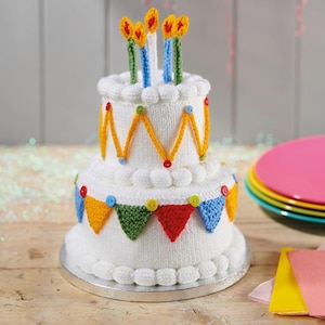 Może przedstawiać: Dwupoziomowy, biały, dziergany tort urodzinowy z kolorowymi akcentami. Ciasto jest ozdobione żółtymi zygzakami, kolorowymi trójkątnymi flagami i zwieńczone świecami. Ciasto stoi na srebrnej paterze.