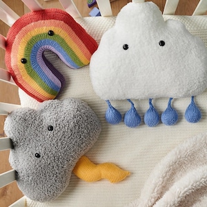 Puede incluir: Tres cojines de peluche con formas meteorológicas: un arcoíris tejido con rayas rojas, naranjas, amarillas, verdes, azules y moradas, una nube blanca esponjosa con cinco gotas de lluvia azules colgantes y una nube gris con un rayo amarillo.