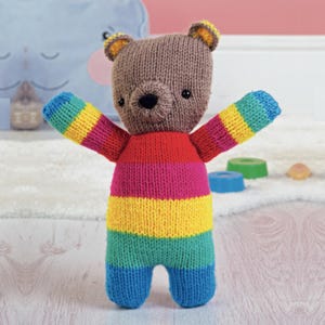 May include: A knitted bear toy with a brown head, black eyes, and a black nose. The bear's body is striped with rainbow colours: red, pink, yellow, green, and blue. The bear's arms are outstretched, and it has blue legs.