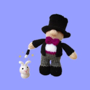 Puede incluir: Muñeco de mago tejido con sombrero de copa negro, esmoquin y pajarita magenta. El muñeco sostiene una varita mágica, conjurando un pequeño conejo blanco con una cara sonriente. El fondo es de color púrpura claro.