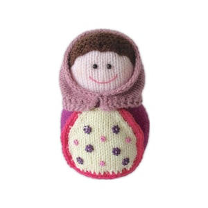 May include: A handmade knitted doll with a smiling face, wearing a pink head covering and a purple and white dress. The dress features a floral design with purple and pink flowers. The doll is crafted with soft yarn and has a charming, whimsical appearance.