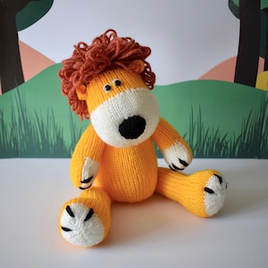 Puede incluir: Un juguete de león tejido a mano en tonos amarillo, blanco y marrón. El león tiene una melena marrón, una cara blanca y una nariz negra. El juguete está sentado con las piernas estiradas, mostrando sus patas con puntas blancas.