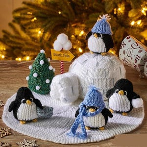 May include: A white knitted igloo with a sign that says "South Pole" and five knitted penguins. The penguins are wearing blue and white hats and scarves. The igloo is sitting on a white knitted surface.
