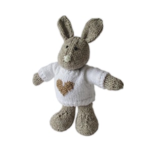 May include: A knitted bunny toy with a white sweater featuring a brown heart. The bunny has gray ears, head, arms, and legs. The toy is made of soft yarn and is designed to be a cuddly companion.