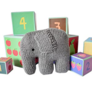 Op de afbeelding: Een grijs gebreid olifantenspeelgoed staat tussen kleurrijke houten blokken. De blokken tonen cijfers en fruitillustraties, waaronder appels en aardbeien. De olifant heeft kleine zwarte ogen en een textuur gebreid ontwerp.