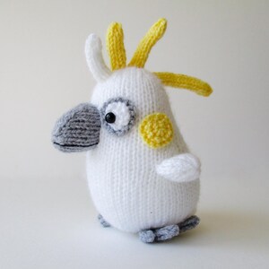 May include: A knitted cockatoo toy featuring a white body, a grey beak, and yellow crest feathers. The bird has a round yellow patch on its cheek, black bead eyes, and small grey feet. The texture is consistent with yarn knitting throughout.